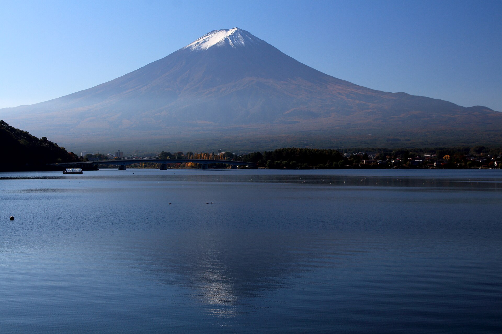 河口湖の湖面越しに見た富士山。湖に架かる橋と対岸の町並みが、山すその手前に並びます