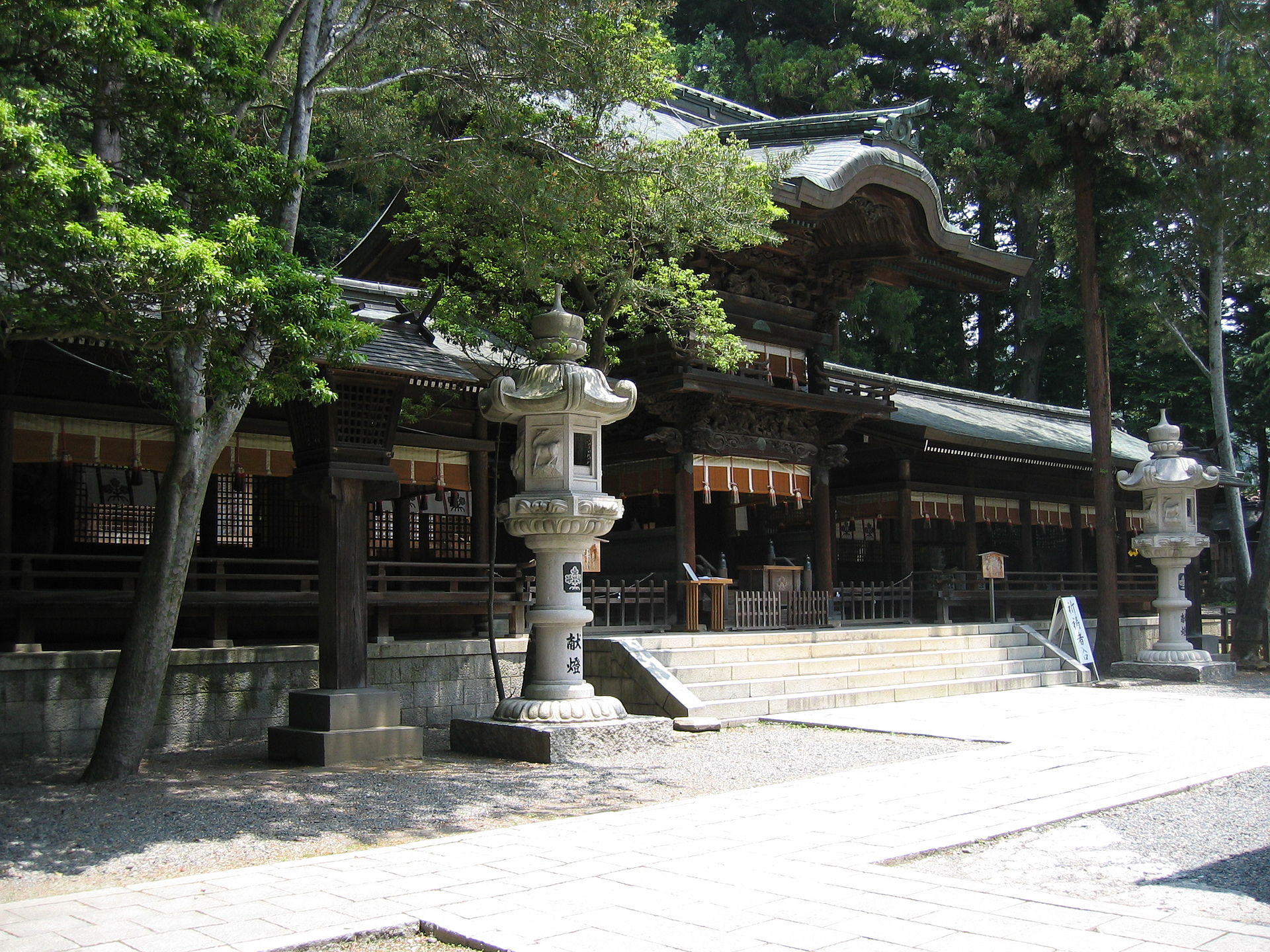 下社秋宮の幣拝殿。石灯籠の向こうに、二層の屋根と、上層にめぐらされた彫刻が見える