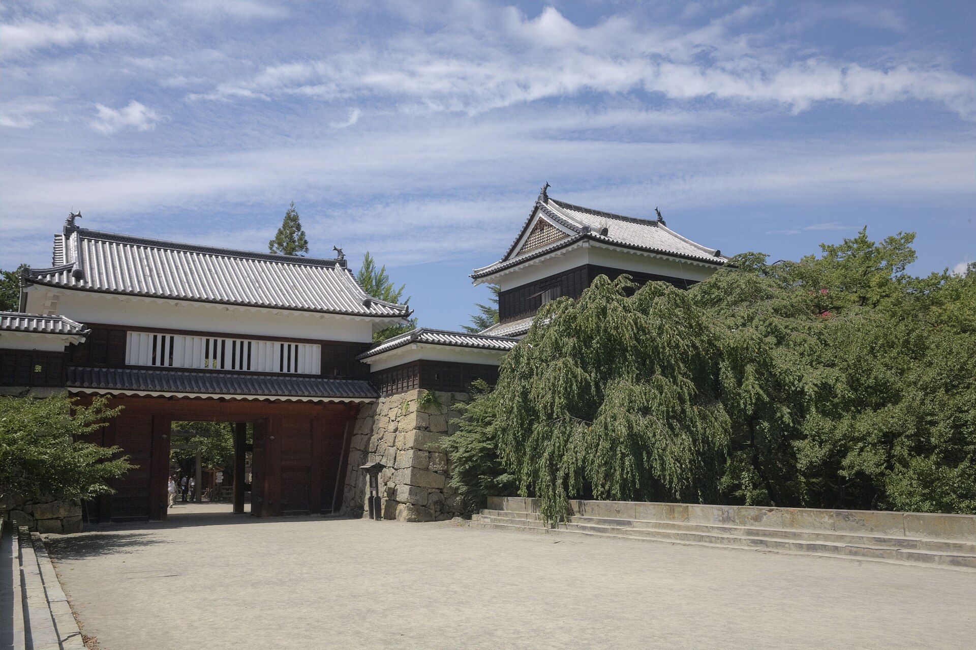 上田城の東虎口櫓門を、砂利敷きの広場側から。門の上に白壁の櫓、右手奥に板壁の櫓と枝垂れの大木